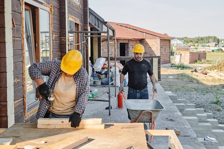 Construction workers building a house, using a saw and carrying materials in a wheelbarrow.