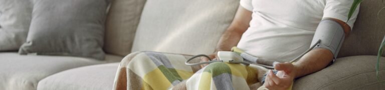 Elderly man monitoring blood pressure on a cozy couch at home, surrounded by medication.