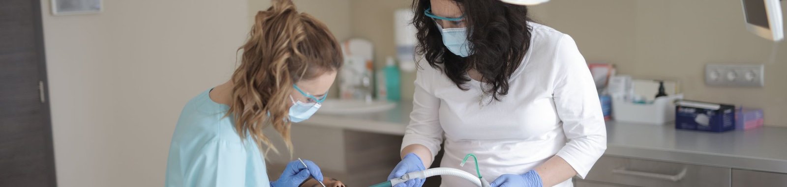Female dentists in medical masks and latex gloves wearing uniform using medical instruments while treating teeth of female patient in modern dental clinic